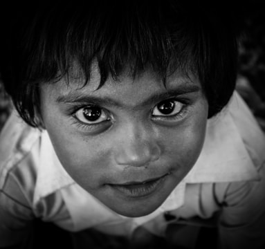 A close-up portrait of a young child with expressive eyes, looking directly at the camera. The child has short, dark hair and is wearing a collared shirt. The image is in black and white, highlighting the contrast and details of the child's facial features.