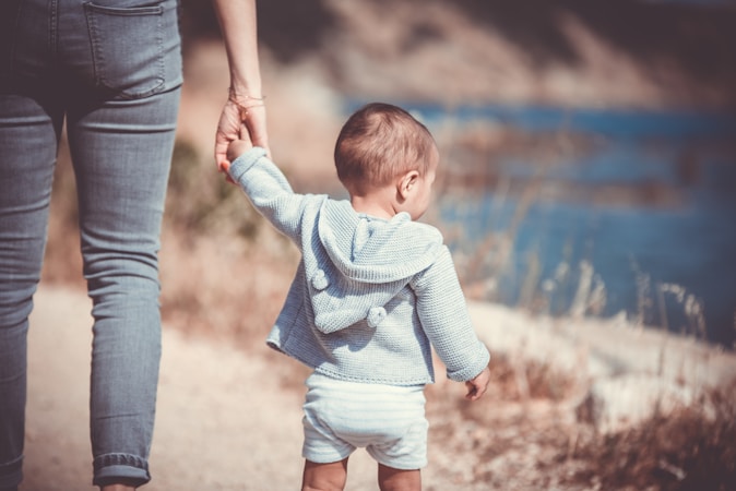 A toddler in a light blue hoodie and white shorts is holding an adult's hand while walking along a path near a body of water. The scene is set outdoors with blurred natural surroundings in the background.