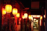 A narrow alleyway adorned with traditional red Chinese lanterns hanging from wooden posts. The warm glow of the lanterns illuminates the rustic walls and lush greenery surrounding the entrance. In the background, a sign with Chinese characters is visible, adding to the cultural atmosphere.