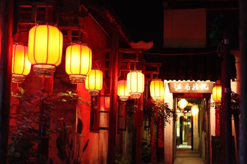 A narrow alleyway adorned with traditional red Chinese lanterns hanging from wooden posts. The warm glow of the lanterns illuminates the rustic walls and lush greenery surrounding the entrance. In the background, a sign with Chinese characters is visible, adding to the cultural atmosphere.