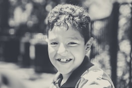 A young child with curly hair smiling broadly, showing a missing tooth. The image is in black and white, with a soft focus background that includes blurred lights and a fence.