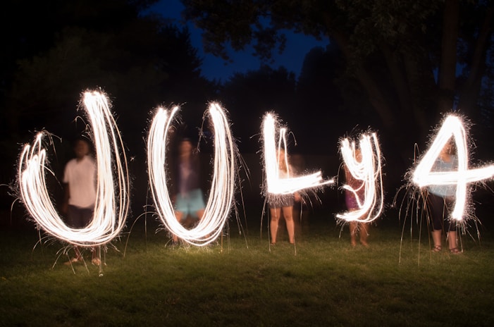 The image features the words 'July 4' written with sparklers in the air, against a dark outdoor background. The scene is illuminated by the light trails, with shadows of people standing in the background holding the sparklers.