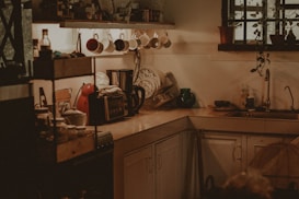 A cozy kitchen corner with dishes drying on a rack, a collection of mugs hanging under a shelf, and a few kitchen appliances on the counter including a kettle and a toaster. Potted plants hang near a window, adding a touch of greenery. Light filters softly into the room, creating a warm and inviting atmosphere.