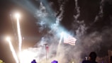 A large American flag is illuminated against a night sky filled with bright fireworks and smoke, creating a vibrant and dynamic scene. The light trails of the fireworks contrast with the dark background, while beams of light and billowing smoke add a dramatic effect.