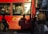 A red city bus features an advertisement for clean air initiatives in London. The bus's side windows reveal passengers inside, some using their phones. Shadows cast by pedestrians are visible on the bus. A person with a hat and carrying a bag is near the bus.