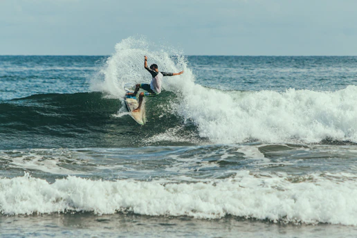 A surfer skillfully rides a wave, creating a dynamic spray of water. The ocean stretches into the distance, with a clear sky above.