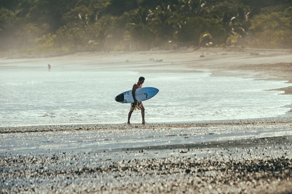 hombre con camisa azul sosteniendo una tabla de surf blanca caminando en la playa durante el día