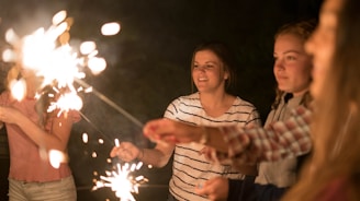 A group of young people holding sparklers in a dimly lit environment, with expressions of joy and celebration. The sparklers are creating bright bursts of light, illuminating their faces and casting a warm glow.