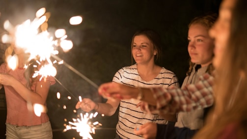 A group of young people holding sparklers in a dimly lit environment, with expressions of joy and celebration. The sparklers are creating bright bursts of light, illuminating their faces and casting a warm glow.