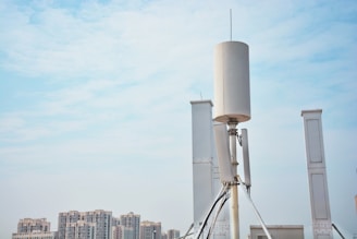 A tall, cylindrical antenna structure is prominently positioned against a clear blue sky. The background features modern high-rise buildings, suggesting an urban setting. The antenna is supported by various cables and metal frames.
