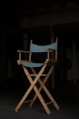 A wooden director's chair with a light blue fabric seat and backrest stands prominently in a dimly lit room. The chair is positioned on a dark floor, with shadows obscuring much of the background, giving the scene an intimate and focused atmosphere.