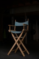 A wooden director's chair with a light blue fabric seat and backrest stands prominently in a dimly lit room. The chair is positioned on a dark floor, with shadows obscuring much of the background, giving the scene an intimate and focused atmosphere.