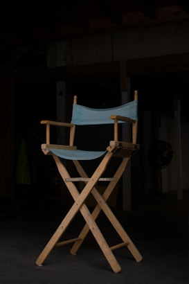 A wooden director's chair with a light blue fabric seat and backrest stands prominently in a dimly lit room. The chair is positioned on a dark floor, with shadows obscuring much of the background, giving the scene an intimate and focused atmosphere.