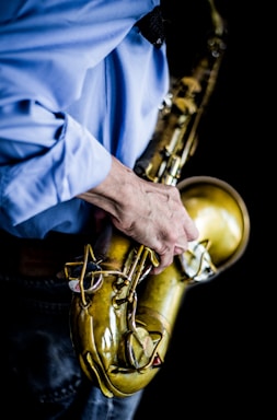 A close-up view of a person wearing a blue shirt holding a brass saxophone. The focus is on the hand gripping the instrument, with emphasis on the details of the saxophone's keys and curves. The background is dark, which makes the metallic finish of the saxophone stand out.