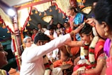 A vibrant and colorful wedding ceremony with a couple seated and surrounded by people, adorned with garlands and traditional attire. The setting includes floral decorations and the guests are engaged in the ritual.