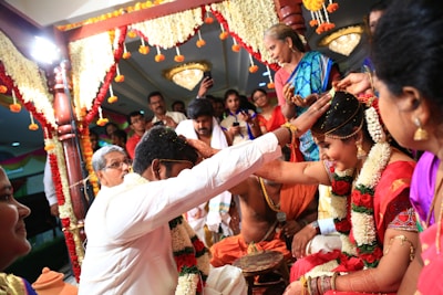 A vibrant and colorful wedding ceremony with a couple seated and surrounded by people, adorned with garlands and traditional attire. The setting includes floral decorations and the guests are engaged in the ritual.