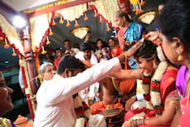 A vibrant and colorful wedding ceremony with a couple seated and surrounded by people, adorned with garlands and traditional attire. The setting includes floral decorations and the guests are engaged in the ritual.