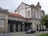 A historic building with arched windows and a sign reading 'VIANA DO CASTELO' on the facade. It features a combination of stone masonry and a red-tiled roof. There are several cars parked in front along a cobblestone street. A leafy tree is visible to the right.