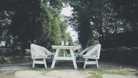 A serene outdoor setting featuring a white wooden picnic table flanked by two matching benches. The furniture is set on a stone patio surrounded by lush green trees, with a stone wall partially visible in the background. The table holds two clear cups, suggesting a peaceful atmosphere for enjoying drinks outside.
