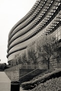 A modern building with a curved facade featuring a grid of windows and external architectural elements. Trees line the base of the structure, which is bordered by a terrace and railing. A paved area and a trash bin are visible in the foreground, completing the urban setting.