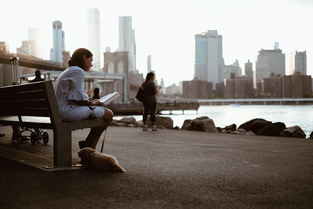 woman sitting on bench reading book with dog during daytime