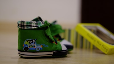 A pair of green baby shoes with a cute animal and vehicle design printed on the side. The shoes have plaid-patterned tops and white soles, with a blurred yellow box in the background.