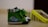 A pair of green baby shoes with a cute animal and vehicle design printed on the side. The shoes have plaid-patterned tops and white soles, with a blurred yellow box in the background.