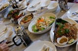 A dining table set with a variety of dishes including a salad with lettuce, eggs, and garnish, a bowl of noodles with herbs, and other various Asian dishes. There are cups filled with drink, a French press for coffee, cutlery, and condiments. The atmosphere appears to be casual dining with a focus on a meal shared among several people.