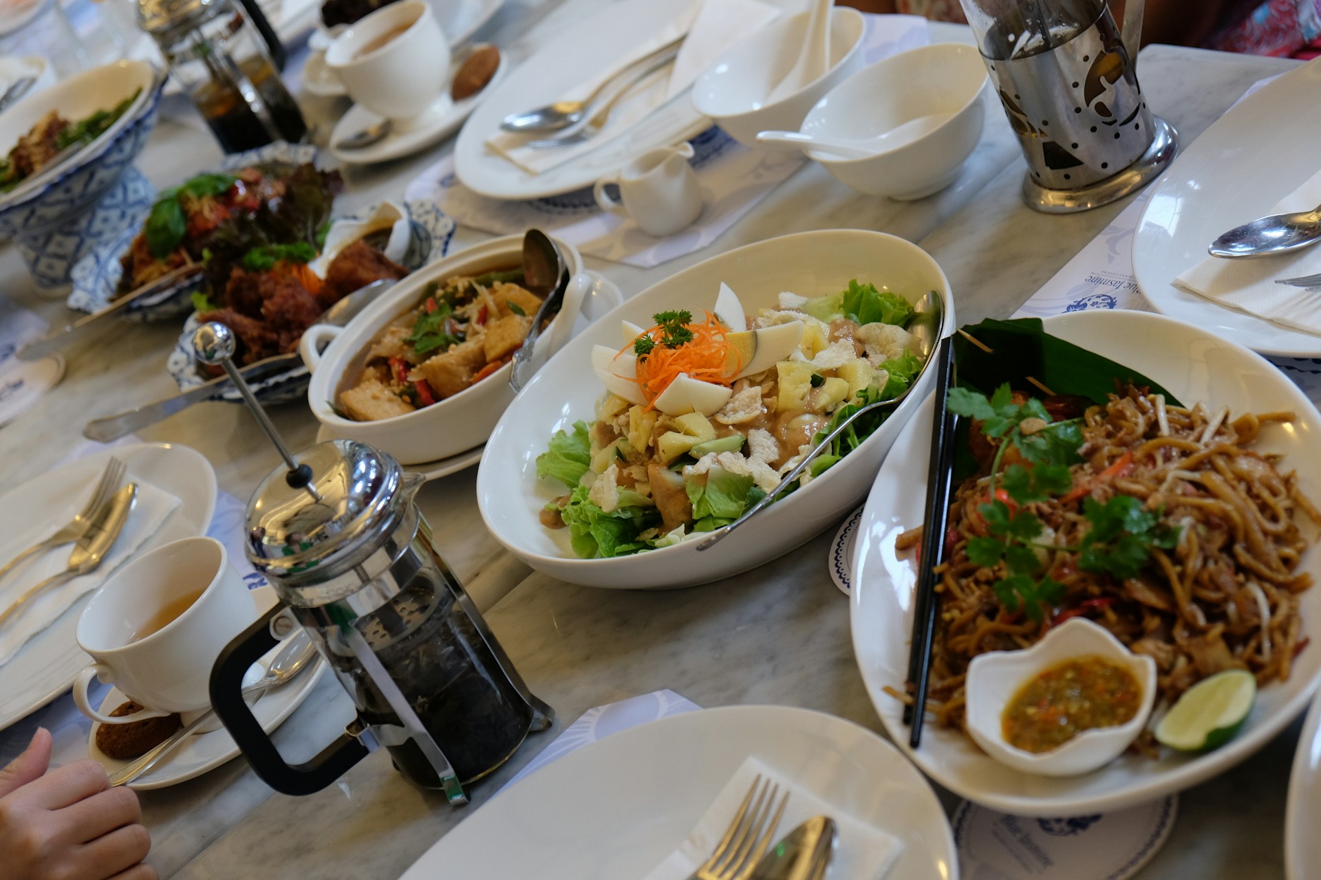 A dining table set with a variety of dishes including a salad with lettuce, eggs, and garnish, a bowl of noodles with herbs, and other various Asian dishes. There are cups filled with drink, a French press for coffee, cutlery, and condiments. The atmosphere appears to be casual dining with a focus on a meal shared among several people.