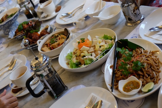 A dining table set with a variety of dishes including a salad with lettuce, eggs, and garnish, a bowl of noodles with herbs, and other various Asian dishes. There are cups filled with drink, a French press for coffee, cutlery, and condiments. The atmosphere appears to be casual dining with a focus on a meal shared among several people.