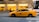 A yellow taxi moves swiftly down a city street, with a blurred background suggesting motion. Buildings and a bus are visible in the backdrop.