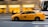 A yellow taxi moves swiftly down a city street, with a blurred background suggesting motion. Buildings and a bus are visible in the backdrop.
