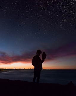 silhouette of man holding hat near body of water during golden hour