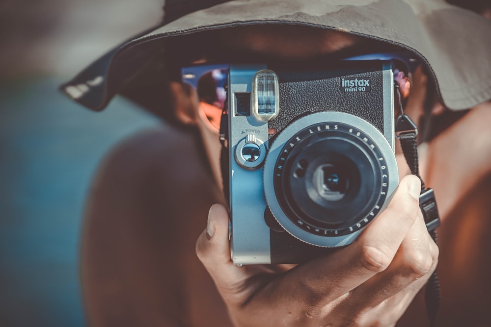 Person mit braunem Fischerhut mit schwarzer und grauer Fujifilm Instax-Kamera