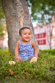 A smiling baby wearing a blue and white patterned outfit sits on lush green grass, leaning against a tree trunk. The background features blurred foliage and a hint of a building, contributing to a vibrant and serene outdoor setting.