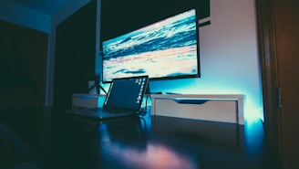 A modern and minimalist workspace featuring a large monitor displaying a scenic ocean wave and sunset image. The setup includes a sleek laptop placed on a dark reflective desk surface, complemented by ambient blue lighting that enhances the futuristic ambiance of the room.