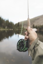 A beginner smiling while holding a hand rod beside a peaceful lake surrounded by trees.