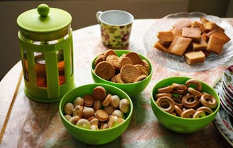 A selection of snacks including various types of cookies and crackers arranged in three green bowls on a table. Nearby, there is a French press with tea, a floral mug, and a stack of plates with floral designs. In the background, there is a plate filled with square-shaped cookies.