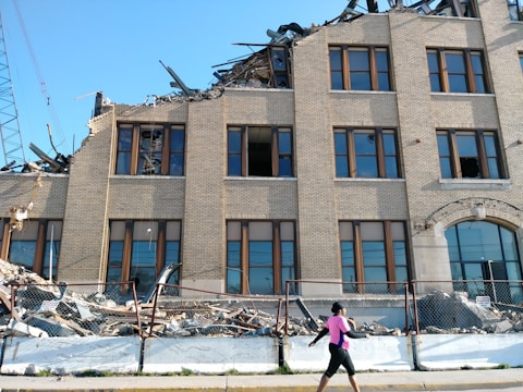 A partially collapsed building with significant structural damage, where the upper section appears destroyed. Bricks and debris are scattered around the base, and some parts of the structure are exposed. A person in athletic clothing is walking in front of the building on a sidewalk. A chain-link fence separates the person from the rubble.