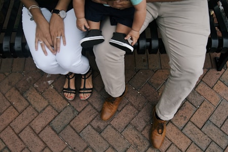 A family setting with two adults and a child sitting on a black metal bench. The adults are wearing white pants and light brown pants, with the person on the left in sandals and the person on the right in brown shoes. The child is sitting on the lap of one adult, wearing black shoes and shorts.