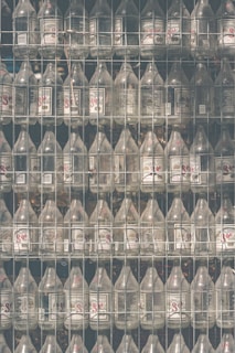 Rows of empty glass bottles are arranged neatly within a metal wire shelving unit, each with a label featuring text and graphics. The setup suggests storage or organization, with light creating a soft reflection on the bottles' surfaces.