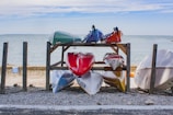 Several kayaks are stored on a wooden rack by the seashore. The rack holds two levels of kayaks, with colorful kayaks on both levels indicating red, white, blue, green, and orange. The background features a calm sea and a partly cloudy sky. The area around the rack is covered in gravel and pebbles.