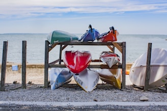 Several kayaks are stored on a wooden rack by the seashore. The rack holds two levels of kayaks, with colorful kayaks on both levels indicating red, white, blue, green, and orange. The background features a calm sea and a partly cloudy sky. The area around the rack is covered in gravel and pebbles.