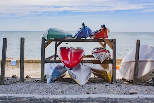 Several kayaks are stored on a wooden rack by the seashore. The rack holds two levels of kayaks, with colorful kayaks on both levels indicating red, white, blue, green, and orange. The background features a calm sea and a partly cloudy sky. The area around the rack is covered in gravel and pebbles.