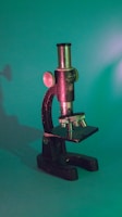 A vintage microscope with a metallic body and knobs, facing slightly to the side, set against a green background. The lighting casts a shadow and adds a soft, warm glow to parts of the microscope, highlighting its intricate details.