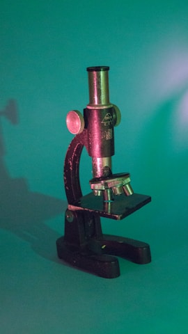 A vintage microscope with a metallic body and knobs, facing slightly to the side, set against a green background. The lighting casts a shadow and adds a soft, warm glow to parts of the microscope, highlighting its intricate details.