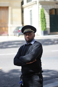A person stands on the street with arms crossed, wearing a black uniform and a cap with 'VOITURIER' written on it. The background includes a building with a wooden door, some greenery, and a sidewalk.