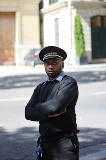 A person stands on the street with arms crossed, wearing a black uniform and a cap with 'VOITURIER' written on it. The background includes a building with a wooden door, some greenery, and a sidewalk.