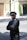 A person stands on the street with arms crossed, wearing a black uniform and a cap with 'VOITURIER' written on it. The background includes a building with a wooden door, some greenery, and a sidewalk.