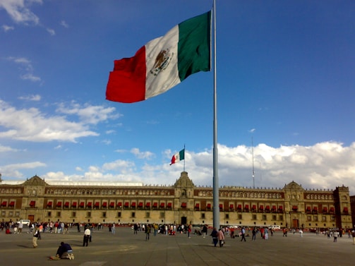 A large Mexican flag waves prominently in the center of a spacious plaza, with a historic building featuring red accents and numerous windows in the background. The sky is bright with scattered clouds, and many people are walking around the open area.